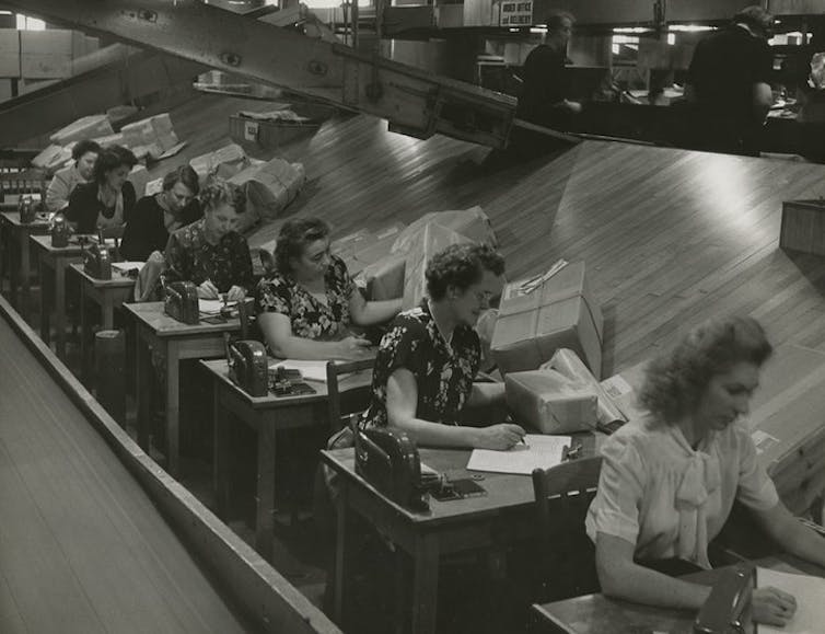 Black-and-white photo of a row of workers sitting at desks with packages.